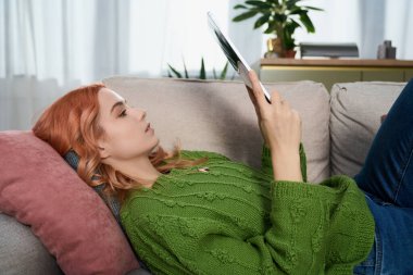 Güzel genç bir kadın güneşli bir günün tadını çıkarırken kendini tabletine kaptırmış bir kanepede dinleniyor..