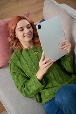 A beautiful young woman relaxes on a sofa, reading on her tablet, surrounded by pillows and comfort.