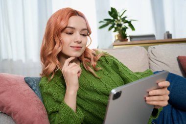 A young woman with beautiful hair relaxes on a sofa while using a tablet in a sunny living room.