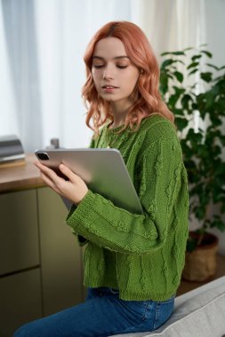 A beautiful young woman with pink hair is engrossed in her tablet while seated in a cozy room.