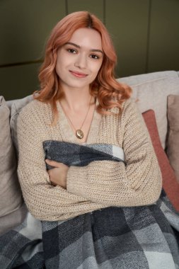 Yumuşak pembe saçlı genç bir kadın kanepeye uzanırken kendini battaniyeye sarar..