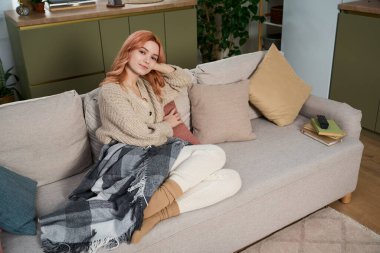 Genç bir kadın sıcak bir battaniyeye sarılı olarak kanepesinde dinleniyor ve huzurlu bir sabahın tadını çıkarıyor..