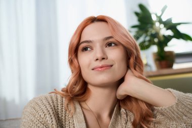 Sevimli genç bir kadın, doğal ışıkla dolu rahat bir yerde rahatlar, memnuniyeti yansıtır..