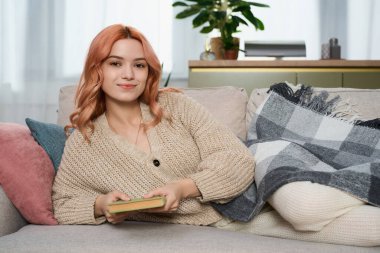 Genç ve güzel bir kadın kanepesinde rahatça uzanırken kitap okumaktan zevk alır..