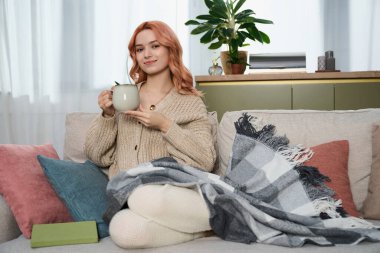 Genç bir kadın, sıcak bir içkiyi sıcak bir battaniyeyle yudumlayarak kanepede dinleniyor..