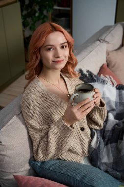 Pembe saçlı genç bir kadın kanepede dinleniyor, kahvesini yudumluyor ve gülümsüyor..