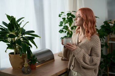 Güzel genç bir kadın bir içki tutuyor, sakin bir bitkinin içindeki pencereden dışarı bakıyor..