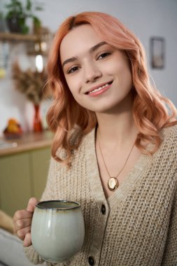 Pembe saçlı genç bir kadın sıcak bir fincan tutarken gülümsüyor, neşe ve rahatlık saçıyor..