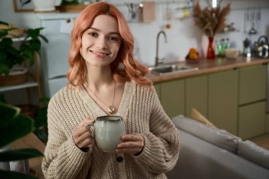 Gülümseyen genç kadın elinde seramik bir kupa tutuyor. Parlak, hoş geldin mutfağında bitkilerle dolu..