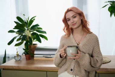 Güzel saçları olan genç bir kadın nazikçe gülümsüyor, elinde parlak, bitki dolu bir mutfakta bir bardak tutuyor..
