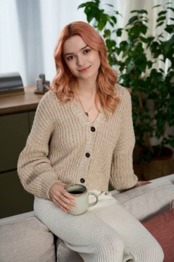 Parlak, davetkar bir odada elinde sıcak bir bardakla gülümseyen pembe saçlı genç bir kadın..