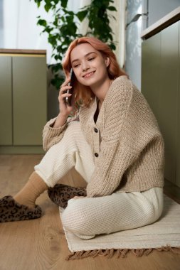 Genç bir kadın, rahat bir atmosferde telefonda konuşurken gülümseyerek yerde oturuyor..