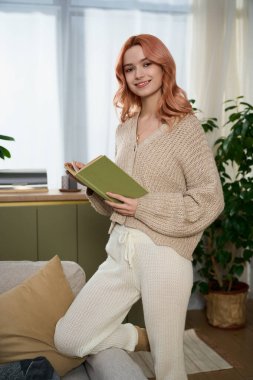 Joyful young woman stands in a warm room, holding a green book with a smile, surrounded by plants.