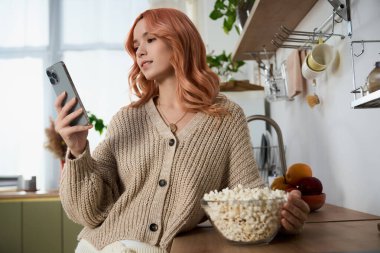 Genç bir kadın parlak bir mutfakta oturmuş elinde patlamış mısırla telefonuna neşeyle bakıyor..