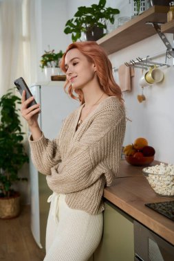 Güzel, genç bir kadın, rahat bir süveter içinde, parlak bir mutfakta dururken telefonuna gülümsüyor..