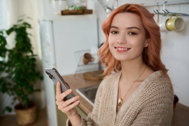 Güzel genç bir kadın, akıllı telefonunu sıcak bir mutfak ortamında tutarken hoş bir şekilde gülümsüyor..