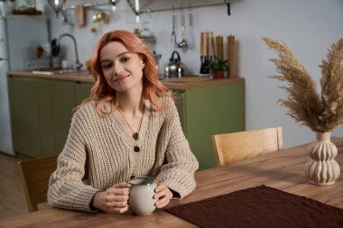 Güzel genç bir kadın ahşap bir masada gevşiyor, şık bir mutfakta sıcak içkisinin tadını çıkarıyor..