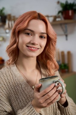 Yumuşak pembe saçlı güzel bir kadın, elinde kupayla gülümsüyor..