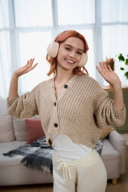 Genç bir kadın, rahat oturma odasında, süveter ve kulaklıkla neşeyle dans ediyor..