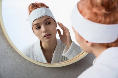 Genç bir kadın yüzüne krem sürüyor, düşünceli bir şekilde aynaya bakıyor..