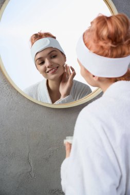 Parlak ve neşeli, genç bir kadın aynada kendine bakarken cilt bakımı yapar..