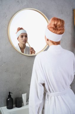 Beyaz cüppeli güzel bir kadın yuvarlak aynaya bakarken cilt bakımı yapıyor..