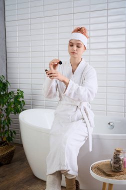 Rahat bir bornoz giymiş genç bir kadın parlak ve huzurlu bir banyoda otururken cilt bakımı yapıyor..