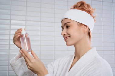 Neşeli genç kadın, huzurlu, aydınlık bir banyoda, kendine bakmanın keyfini süren cilt bakımı serumuna hayran..