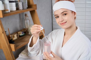 Güzel genç bir kadın, şık banyo düzeninde gülümseyen bir cilt serumu çıkarıyor..