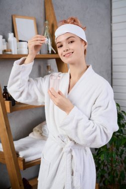 Yumuşak bornozlu genç kadın spa 'sında cilt malzemelerini karıştırırken gülümsüyor..