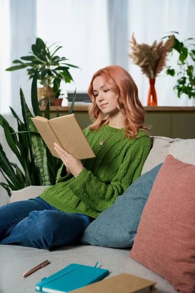 A beautiful young woman enjoys her time reading on a comfortable sofa surrounded by greenery.