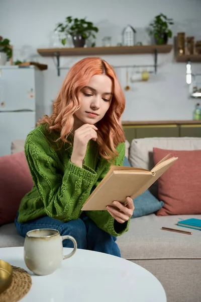 Pembe saçlı genç bir kadın çay ve rahat bir ortamda kitap okuyarak rahatlıyor..