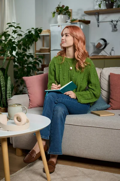 Genç bir kadın, yemyeşil bitkilerin arasında bir not defterine yazarak kanepede dinleniyor..