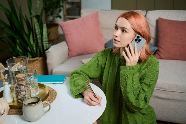 Canlı saçlı genç bir kadın telefonda konuşuyor, rahat bir oturma odasına notlar yazıyor..