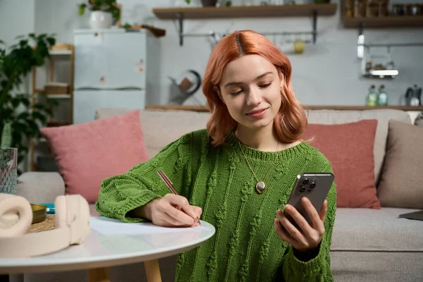 Pembe saçlı güzel bir kadın telefonuna odaklanıyor ve küçük bir masaya notlar yazıyor..