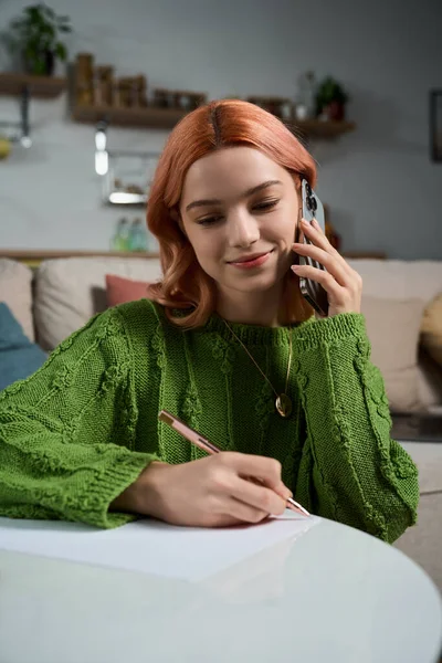 Genç bir kadın, sıcak, davetkar bir odada not defterine yazarken kulağına bir telefon tutuyor..
