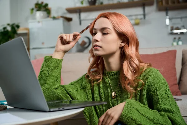 Yeşil kazaklı genç bir kadın sıcak, davetkar bir mekanda dizüstü bilgisayarına dikkatle bakar..