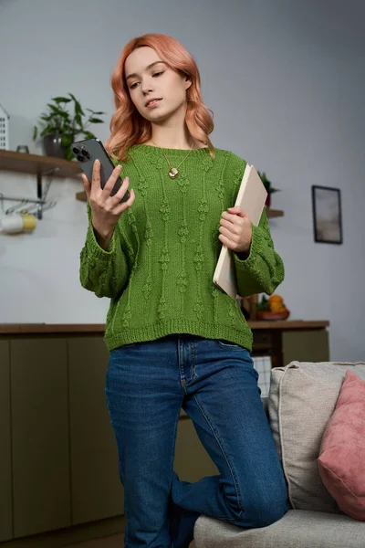 Güzel genç bir kadın yeşil bir kazak giymiş, rahat bir ev ortamında telefonuna bakıyor..