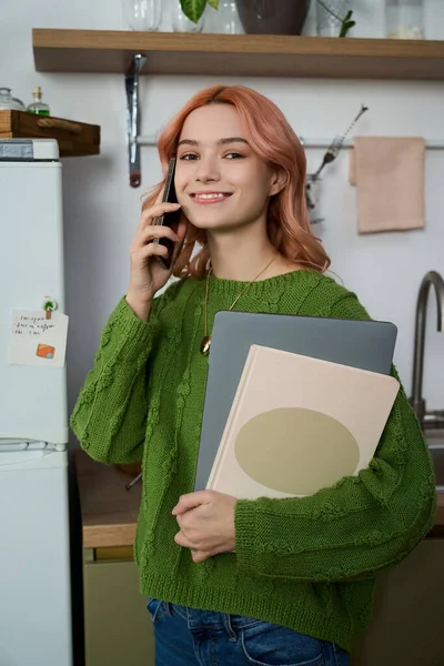 Neşeli genç bir kadın sıcak bir mutfakta telefonuyla konuşurken dizüstü bilgisayar ve defter tutuyor..