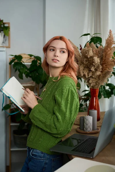 Pembe saçlı güzel genç bir kadın parlak, bitkilerle dolu bir odada deftere yazıyor..