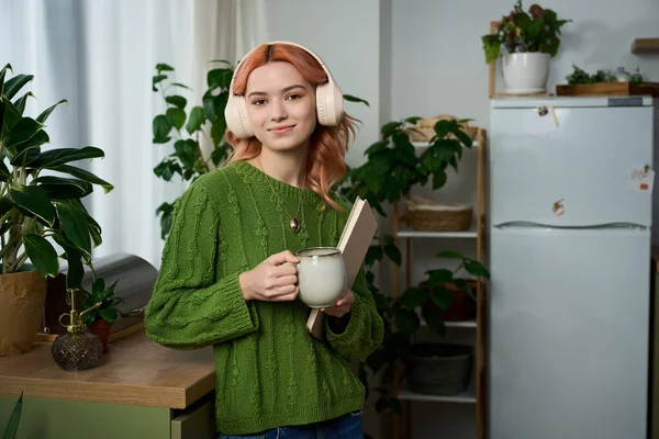 Pembe saçlı genç bir kadın yeşil bir kazakla rahatlıyor, canlı bitkilerin arasında çayının tadını çıkarıyor..
