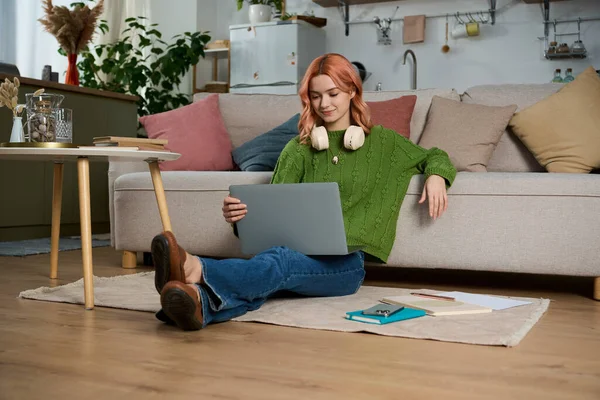 Yeşil kazaklı ve kulaklıklı mutlu genç bir kadın kitapların ve bitkilerin arasında dizüstü bilgisayarının keyfini çıkarıyor..