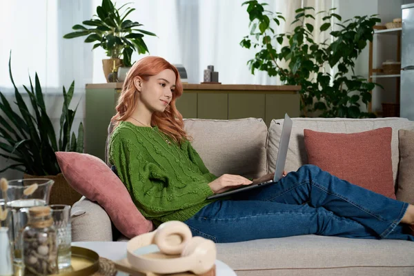 Pembe saçlı güzel bir kadın kanepede rahat rahat dizüstü bilgisayarıyla çalışıyor..