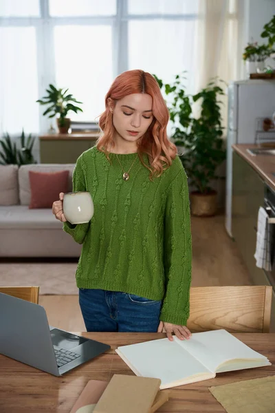 Yeşil kazaklı genç bir kadın parlak bir mutfakta çay yudumluyor, düşüncelere dalmış..