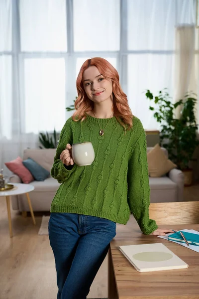 Yeşil kazaklı güzel genç bir kadın güneşli bir odada dinlenirken çay içiyor..