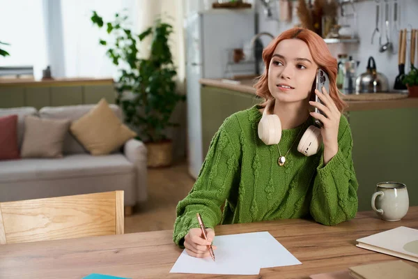 Genç bir kadın, şık bir mutfaktaki ahşap bir masada notlar alırken telefonla konuşuyor..