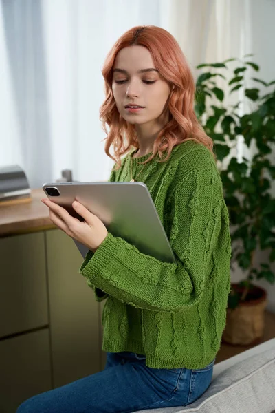 A beautiful young woman with pink hair is engrossed in her tablet while seated in a cozy room.