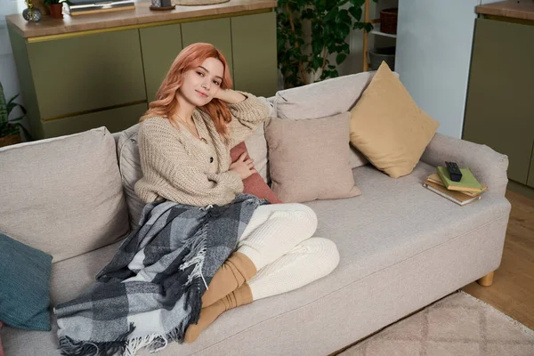 Genç bir kadın sıcak bir battaniyeye sarılı olarak kanepesinde dinleniyor ve huzurlu bir sabahın tadını çıkarıyor..