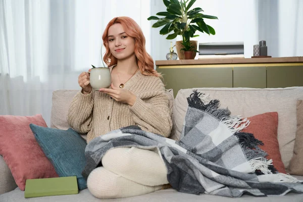 Genç bir kadın, sıcak bir içkiyi sıcak bir battaniyeyle yudumlayarak kanepede dinleniyor..