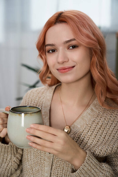 A young woman with soft pink hair smiles, holding a warm drink in a serene setting.